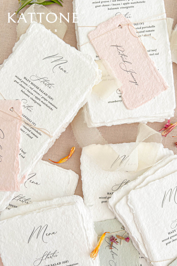 Angled overhead photograph of personalized handmade paper wedding menus arranged with blush pink and sage green place cards. The ivory-white deckled edge menus feature delicate black calligraphy, traditional serif lettering, and individual dinner courses. A blush pink guest name tag attached with natural twine, soft ivory chiffon ribbon, and scattered colorful petals highlight the romantic handmade wedding stationery details.
