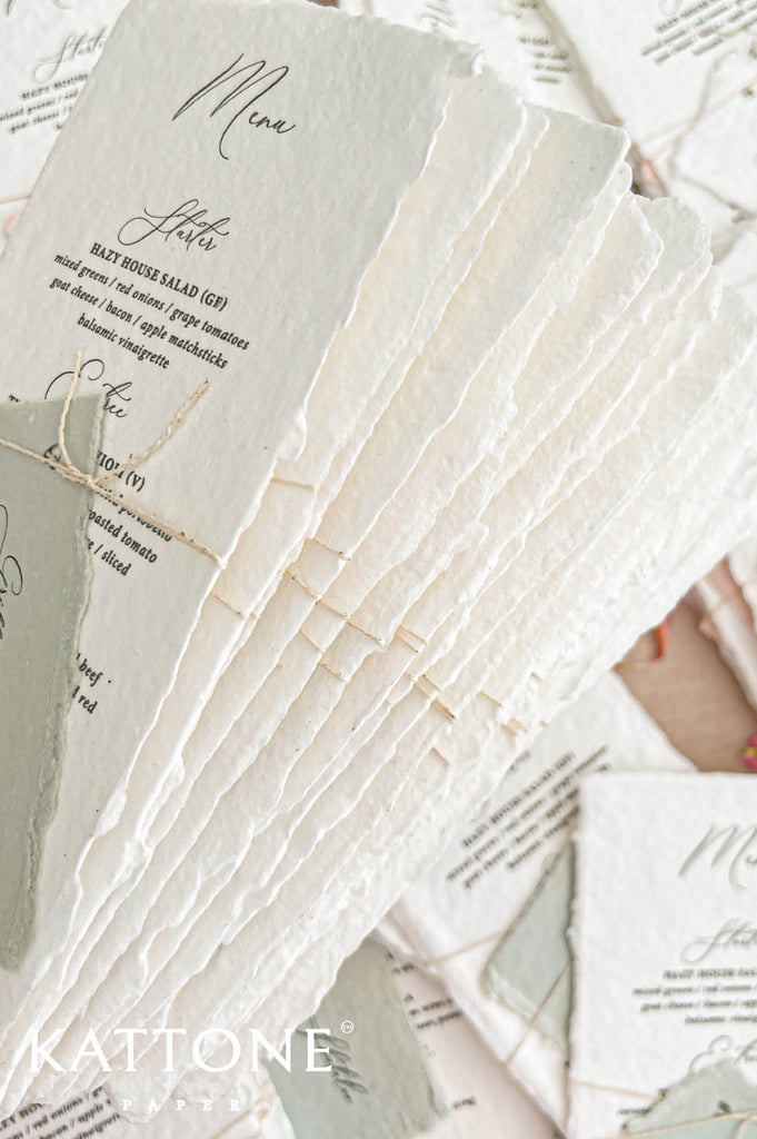 Close-up photograph of a stacked collection of luxury ivory-white handmade paper wedding menus, highlighting their thick textured paper and naturally torn deckled edges. The top menu features black calligraphy, elegant printed dinner courses, and a muted sage green personalized place card secured with natural twine. Coordinating wedding menus are softly visible in the background.