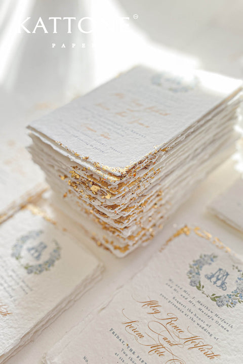 This handmade paper wedding invitation suite combines fine craftsmanship with opulent detailing. Printed on luxury cotton paper with deckled edges, each invitation is accented with subtle touches of gold leaf, catching the light with a refined shimmer that speaks of romance and sophistication. The design features a hand-painted floral monogram, symbolizing the couple’s union, surrounded by soft watercolor greenery and pastel hues.