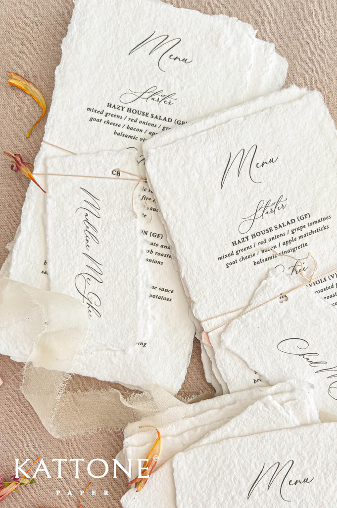 Overhead close-up of elegant ivory-white handmade paper wedding menus featuring textured deckled edges, black calligraphy, and classic serif typography. The menus are paired with personalized handmade paper place cards, secured with delicate natural twine. Soft blush pink and sage green accents, ivory chiffon ribbon, and scattered flower petals complete the romantic wedding stationery arrangement on a beige linen background.