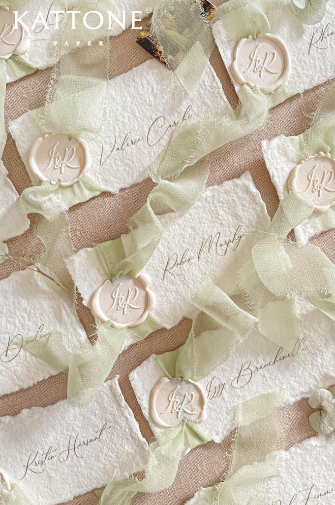 Luxury handmade paper wedding place cards featuring deckled edge cotton paper, elegant calligraphy guest names, sage chiffon ribbon, and wax seal detailing.