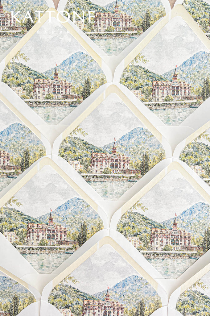 Overhead arrangement of elegant white wedding envelopes featuring illustrated watercolor liners with an Alpine mountain landscape, historic waterfront hotel, peaceful lake, and surrounding greenery. The repeating scenic design, soft ivory envelope borders, and delicate hand-painted details create a coordinated collection of luxury destination wedding envelopes for an elegant European lakeside wedding.