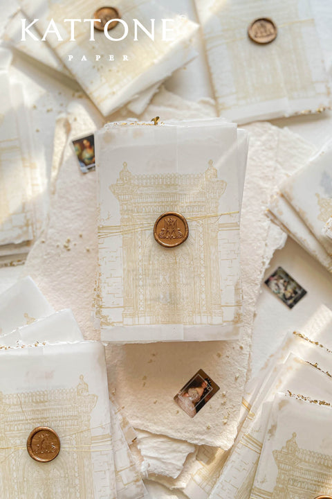 This handmade paper wedding invitation suite combines fine craftsmanship with opulent detailing. Printed on luxury cotton paper with deckled edges, each invitation is accented with subtle touches of gold leaf, catching the light with a refined shimmer that speaks of romance and sophistication. The design features a hand-painted floral monogram, symbolizing the couple’s union, surrounded by soft watercolor greenery and pastel hues.