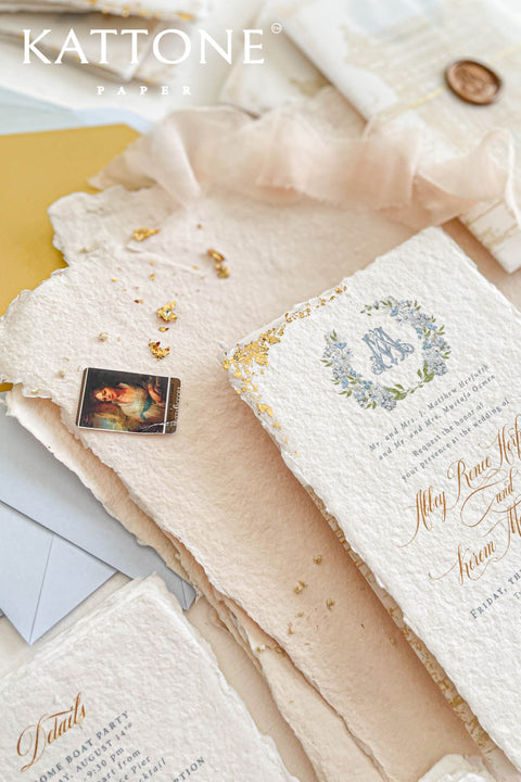This handmade paper wedding invitation suite combines fine craftsmanship with opulent detailing. Printed on luxury cotton paper with deckled edges, each invitation is accented with subtle touches of gold leaf, catching the light with a refined shimmer that speaks of romance and sophistication. The design features a hand-painted floral monogram, symbolizing the couple’s union, surrounded by soft watercolor greenery and pastel hues.