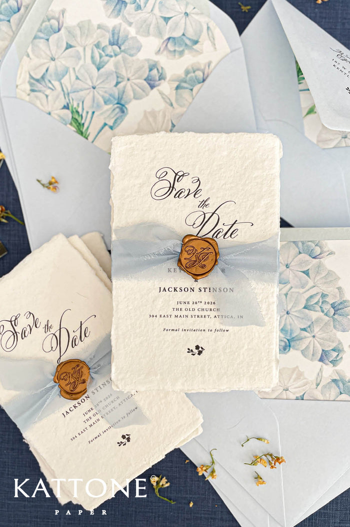 Handmade save the date card on deckled edge cotton paper with soft blue chiffon ribbon, gold wax seal monogram, elegant calligraphy typography and light blue envelope liner with watercolor floral pattern.