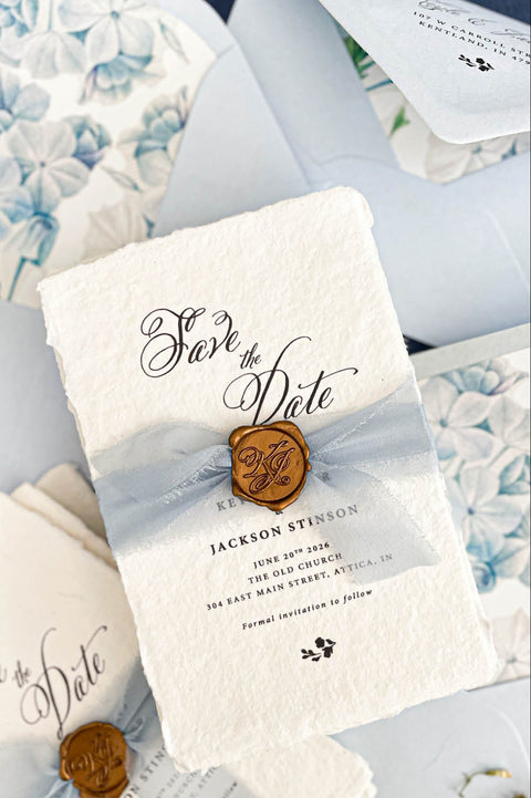 Close up of gold wax seal with custom monogram initials placed over sheer dusty blue chiffon ribbon on textured deckled edge handmade paper card.