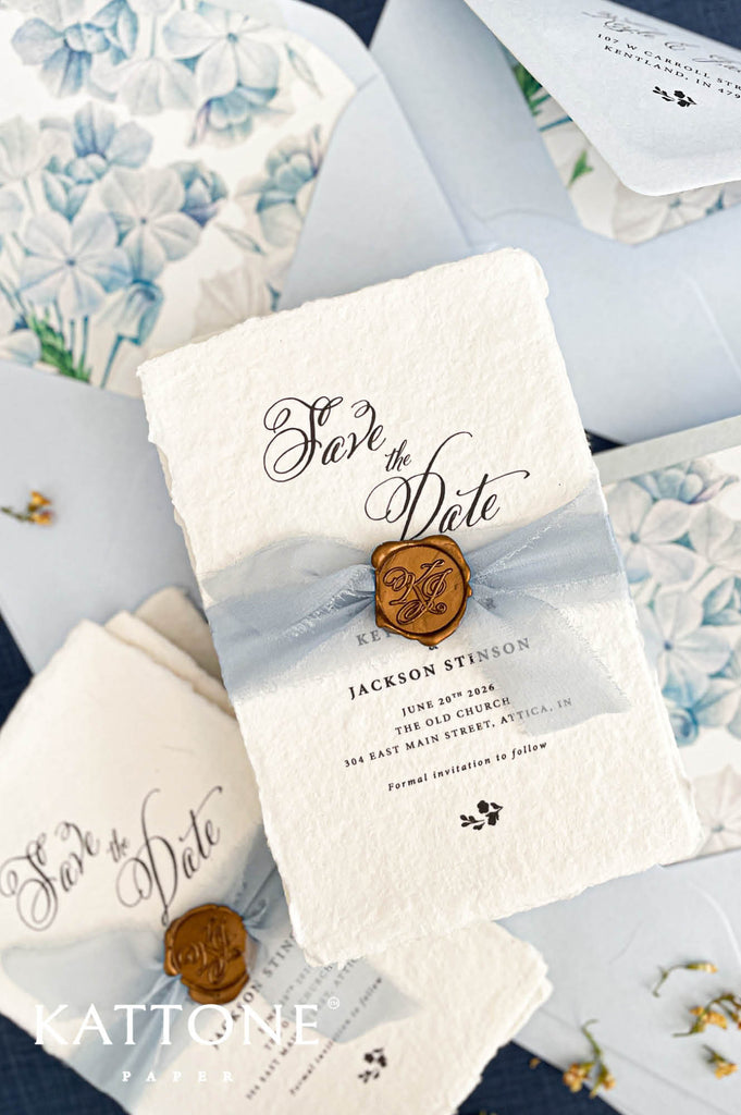 Close up of gold wax seal with custom monogram initials placed over sheer dusty blue chiffon ribbon on textured deckled edge handmade paper card.