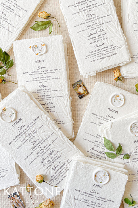Elevate your wedding table with our embossed handmade paper menus, where timeless craftsmanship meets modern refinement. Each menu is printed on 100% cotton handmade paper with delicate deckled edges and a subtle botanical embossing, creating a tactile experience of understated luxury. Finished with an elegant floral wax seal personalized for each guest, these menus add an exquisite fine-art touch to your reception decor.