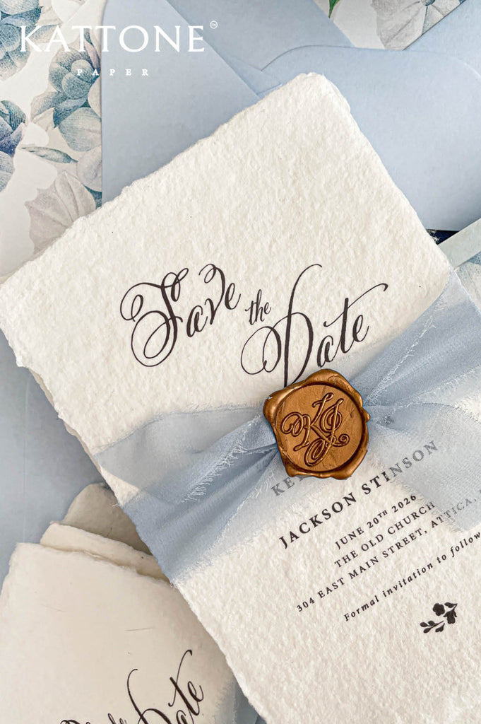 Close up of gold wax seal with custom monogram initials placed over sheer dusty blue chiffon ribbon on textured deckled edge handmade paper card.