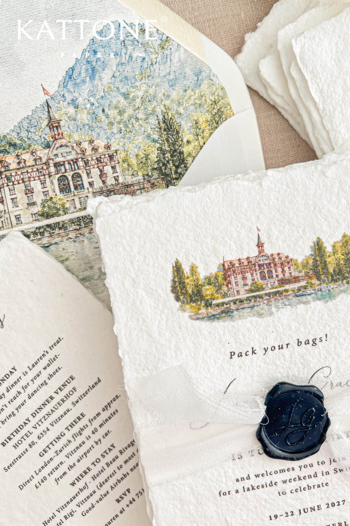 Close-up of an elegant destination birthday invitation on textured ivory handmade paper with deckled edges and a custom watercolor illustration of a Swiss lakeside hotel surrounded by trees and mountains. A delicate ivory ribbon with a personalized navy blue wax seal decorates the invitation beside an open envelope featuring a matching watercolor landscape liner.