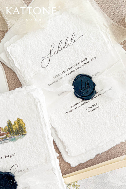 Detailed overhead photograph of a luxury birthday celebration schedule printed on ivory handmade paper with organic deckled edges. Elegant black calligraphy, classic typography, a sheer ivory ribbon and a personalized navy blue wax seal complete the design, accompanied by coordinating watercolor invitation cards and an illustrated Swiss lakeside envelope liner on natural beige linen.