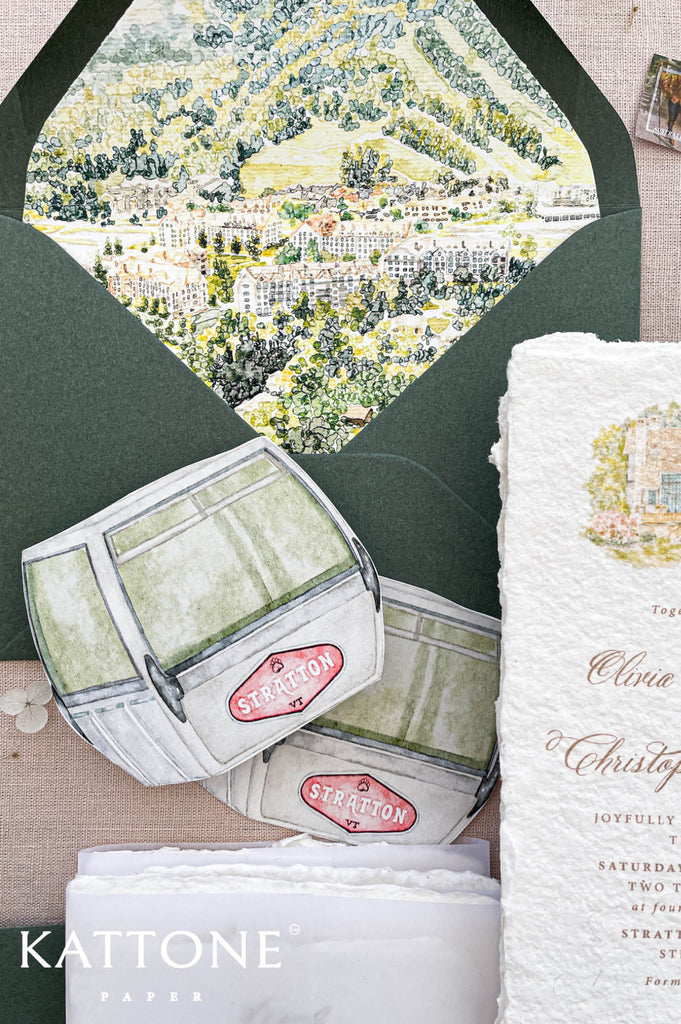 Romantic flat lay of luxury wedding stationery showcasing handmade paper invitations, custom watercolor artwork, scenic vellum wraps, sage green envelopes, details cards, and coordinating wedding inserts.