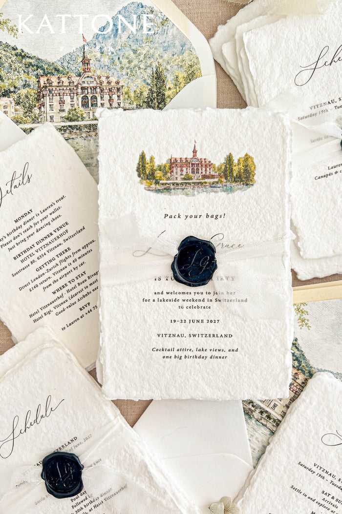 Styled overhead photograph of luxury handmade paper birthday invitations for a Swiss lakeside celebration. The ivory invitation features custom watercolor artwork of a historic waterfront hotel, an ivory ribbon and navy blue personalized wax seal, surrounded by matching schedule and details cards, watercolor envelope liners illustrating Lake Lucerne and the Swiss Alps, and soft beige linen fabric.