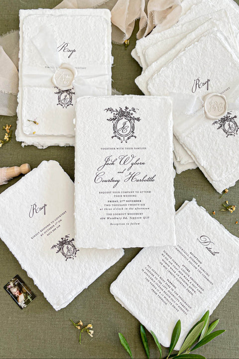 Luxury handmade cotton paper wedding invitation with deckled edges and ornate monogram crest, styled with botanical watercolor envelope liners and coordinated stationery suite.
