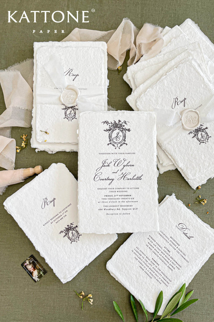 Luxury handmade cotton paper wedding invitation with deckled edges and ornate monogram crest, styled with botanical watercolor envelope liners and coordinated stationery suite.