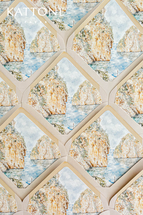 Overhead flat lay of luxury ivory and blush wedding envelopes arranged in a repeating pattern, each featuring an illustrated watercolor liner inspired by Sicily's Faraglioni of Scopello. The scenic Mediterranean artwork depicts golden coastal cliffs, turquoise-blue water, and delicate sage greenery, framed by soft neutral envelope borders for romantic Italian destination wedding stationery.