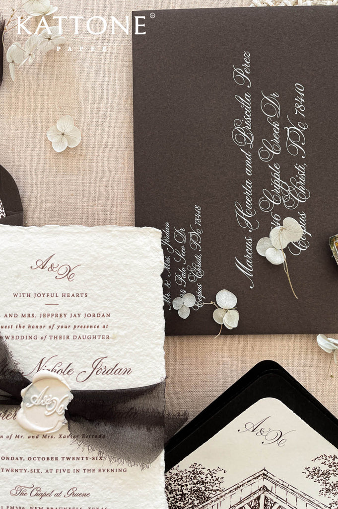 Wedding stationery arranged in a flat lay showcasing handmade paper invitations, dark envelopes, ribbon, wax seal, and chapel artwork.
