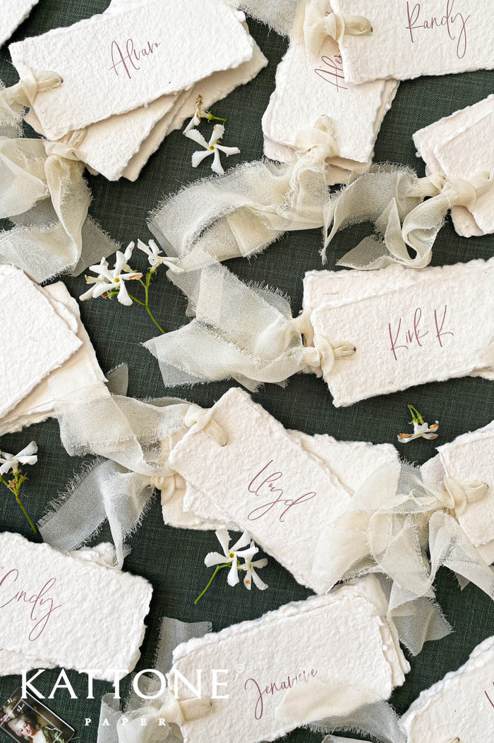 Ivory handmade paper wedding place cards featuring soft deckled edges, elegant calligraphy guest names, and delicate chiffon ribbon ties, styled on a textured green linen background for a romantic tablescape.