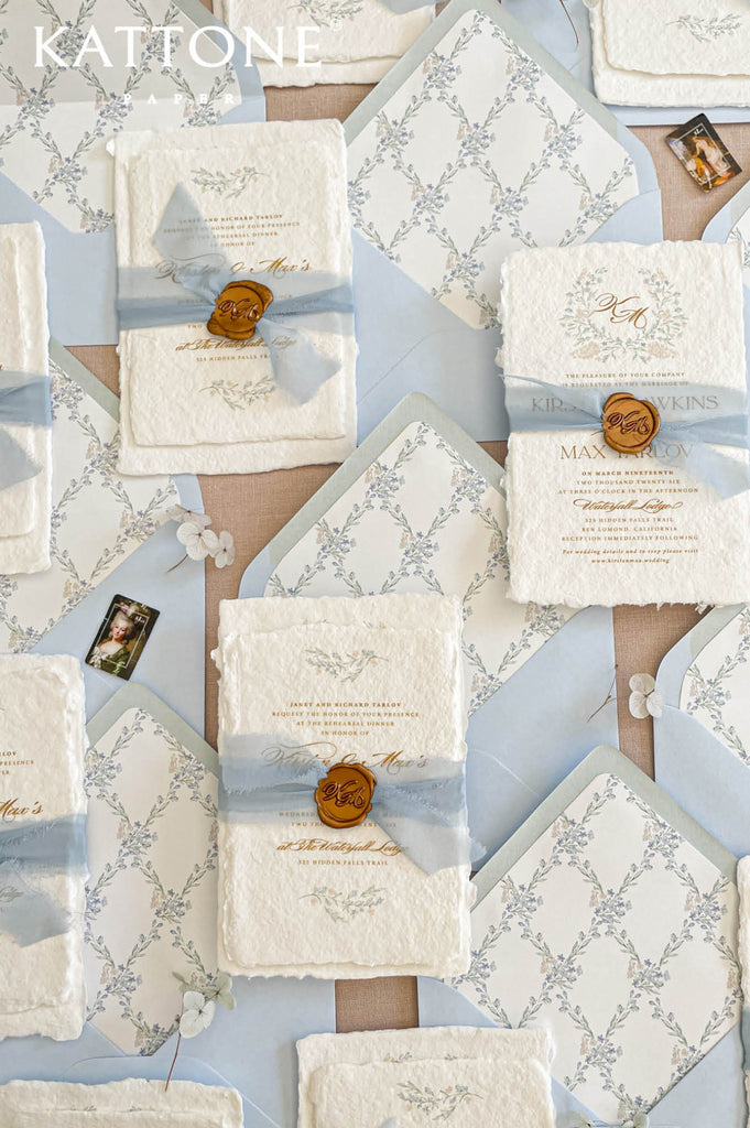 Fine art wedding invitation suite styled with handmade paper invitations, silk ribbon, wax seals, pale blue envelopes and botanical envelope liners on neutral linen background.