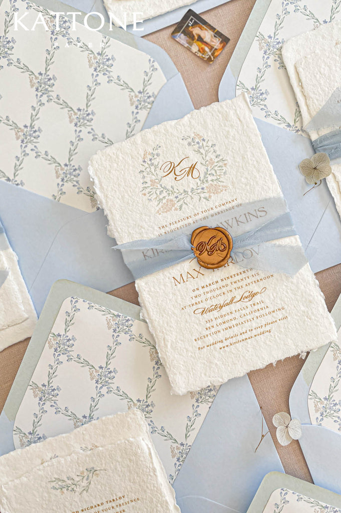 Golden wax seal monogram placed over soft blue silk ribbon on deckled edge handmade paper invitation, highlighting luxury finishing details.