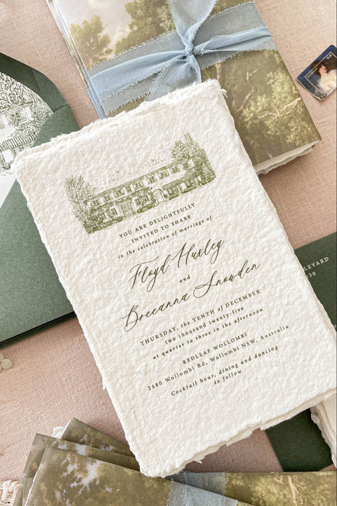 Romantic flat lay of luxury wedding stationery showcasing handmade paper invitations, scenic vellum wraps, custom venue artwork, sage envelopes, details cards, and ribbon styling.