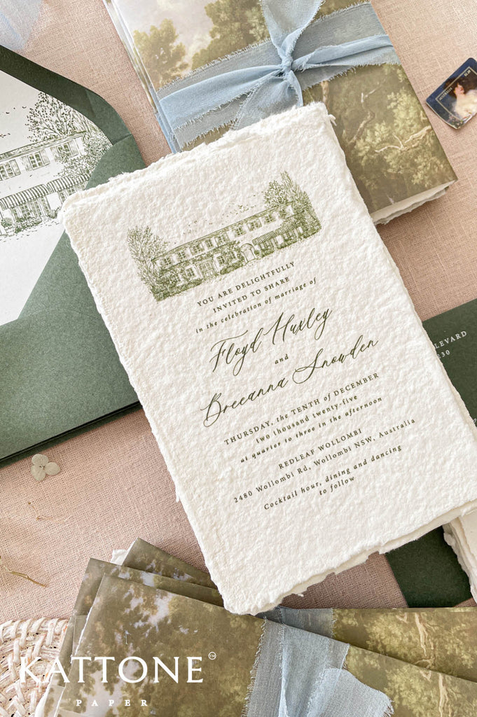 Romantic flat lay of luxury wedding stationery showcasing handmade paper invitations, scenic vellum wraps, custom venue artwork, sage envelopes, details cards, and ribbon styling.