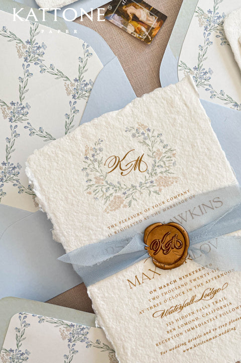 Close up of textured handmade cotton paper wedding invitation showing organic deckled edges, delicate watercolor botanical wreath and elegant gold calligraphy typography.
