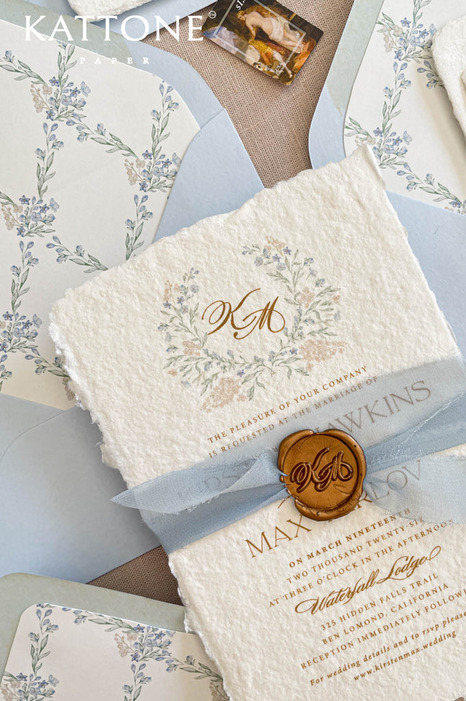 Close up of textured handmade cotton paper wedding invitation showing organic deckled edges, delicate watercolor botanical wreath and elegant gold calligraphy typography.