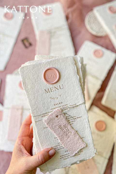 These handmade paper wedding menus capture the essence of romance and artisanal beauty. Crafted from 100% cotton rag paper, each menu features deckled edges, a luxurious tactile finish, and a timeless aesthetic that perfectly complements any elegant tablescape. The menus are adorned with a personalized wax seal and a custom name tag, adding a thoughtful, bespoke touch that enhances your guests’ dining experience.