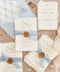 Luxury handmade cotton paper wedding invitation with deckled edges, botanical wreath monogram, soft blue silk ribbon and gold wax seal, styled with coordinating pale blue envelopes and floral liners.