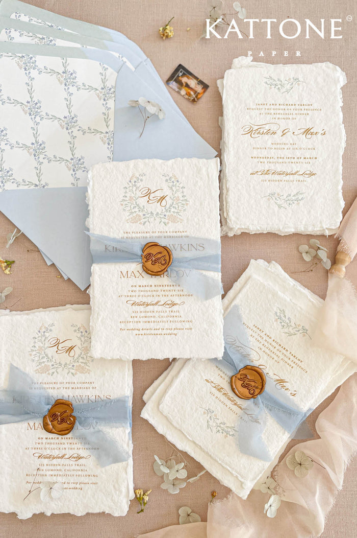 Luxury handmade cotton paper wedding invitation with deckled edges, botanical wreath monogram, soft blue silk ribbon and gold wax seal, styled with coordinating pale blue envelopes and floral liners.