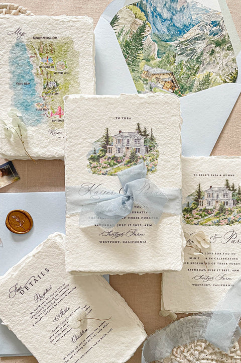 Luxury handmade paper wedding invitation suite featuring custom watercolor venue artwork, scenic envelope liners, watercolor map inserts, deckled edge cotton paper cards, and dusty blue chiffon ribbon styling.