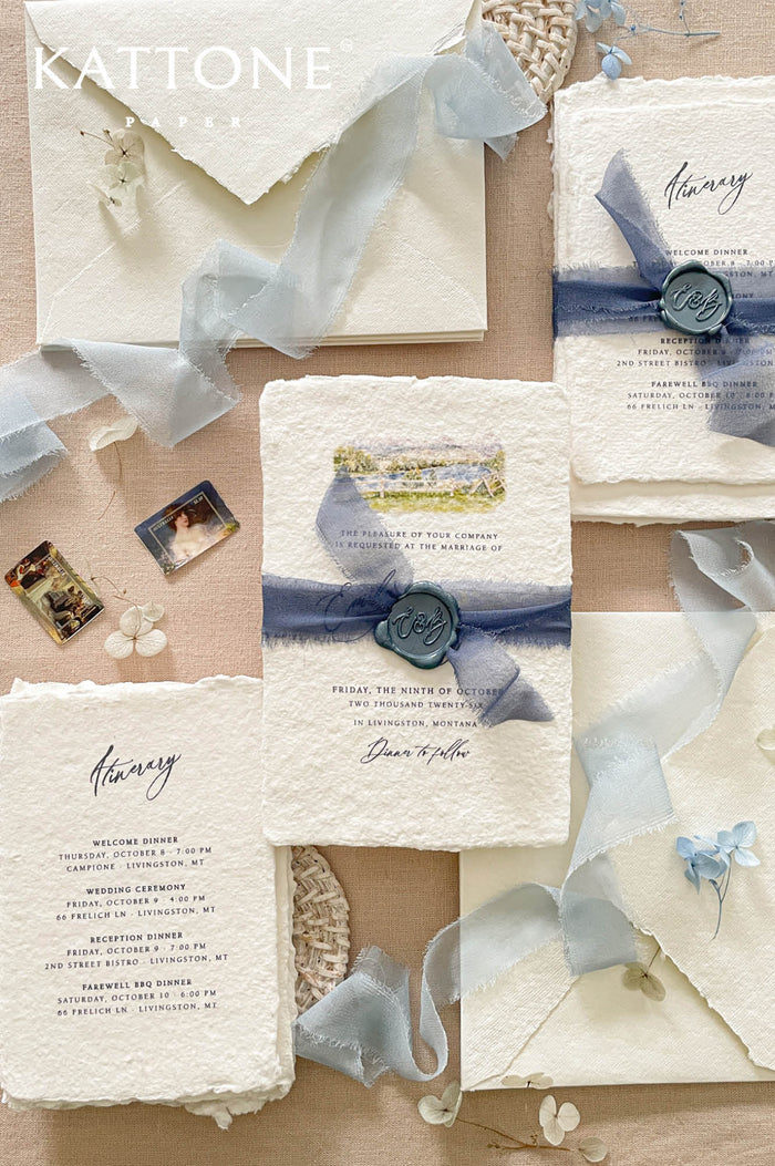 Luxury handmade paper wedding invitation suite featuring deckled edge cotton paper, custom watercolor venue artwork, dusty blue chiffon ribbon wraps, monogram wax seals, itinerary cards, and coordinated ivory envelopes.