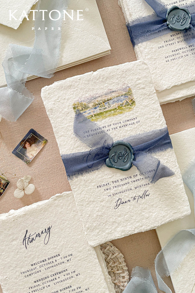 Elegant handmade cotton paper wedding invitation with deckled edges, custom watercolor venue illustration, dusty blue chiffon ribbon wrap, and personalized monogram wax seal.