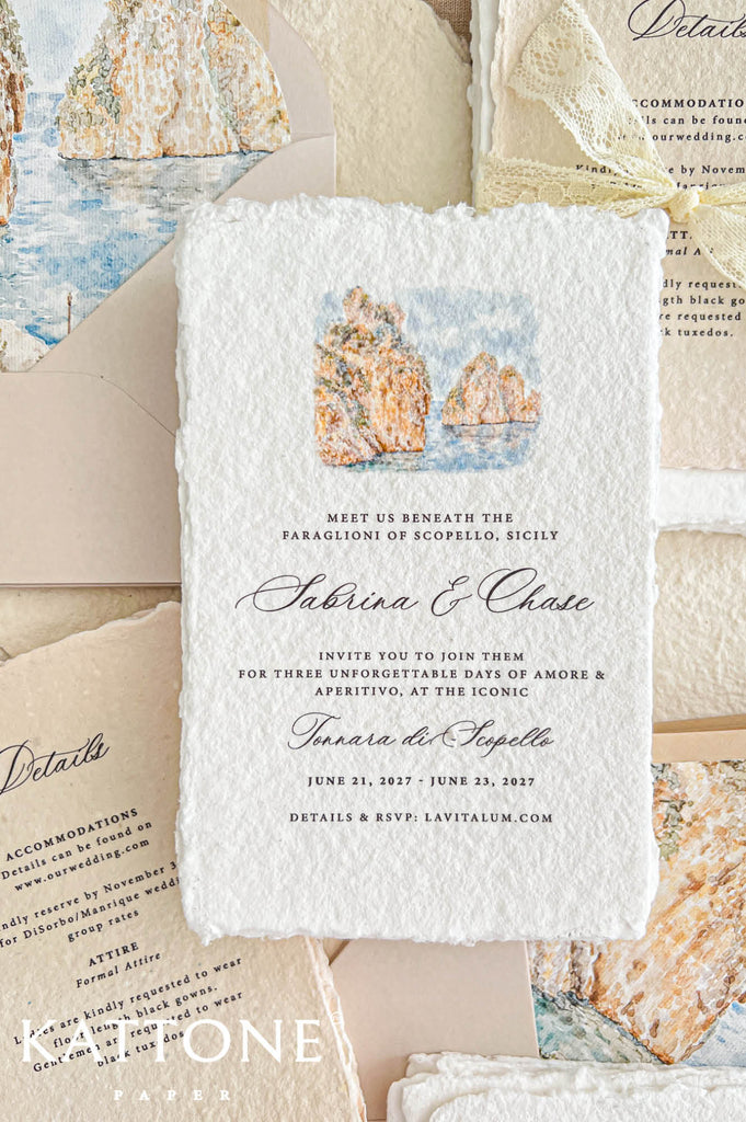 Close-up of a luxury ivory handmade paper wedding invitation featuring a delicate watercolor illustration of the golden limestone cliffs and blue Mediterranean waters of Scopello, Sicily. Elegant black calligraphy and refined typography complement the natural deckled edges, with a champagne envelope and matching coastal watercolor liner, lace-wrapped details cards, and textured handmade paper in the background.