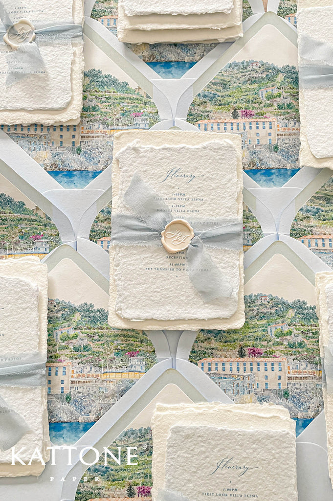 Elegant handmade cotton paper wedding invitation with deckled edges, custom watercolor venue illustration, dusty blue chiffon ribbon wrap, and personalized wax seal in soft coastal tones.