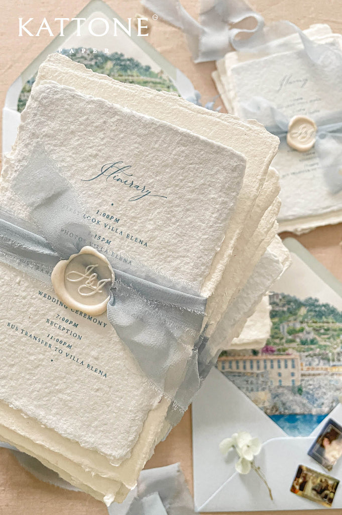 Elegant handmade cotton paper wedding invitation with deckled edges, custom watercolor venue illustration, dusty blue chiffon ribbon wrap, and personalized wax seal in soft coastal tones.