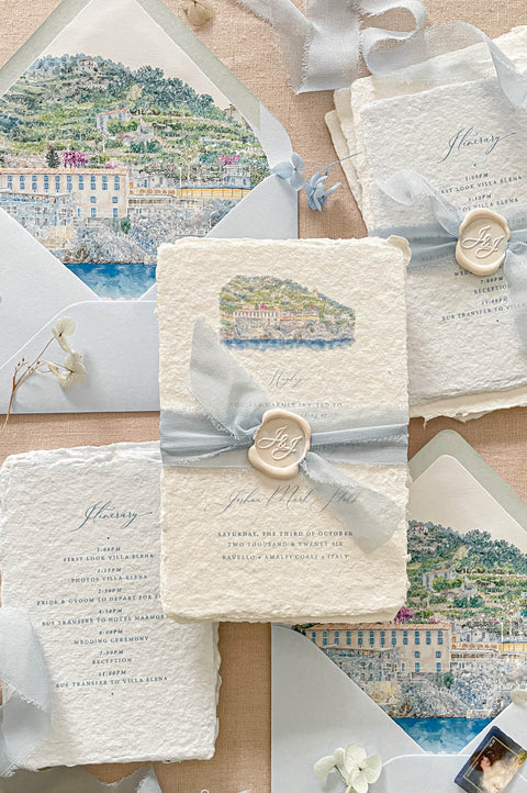 Luxury handmade paper wedding invitation suite featuring deckled edge cotton paper, custom watercolor Amalfi Coast venue artwork, dusty blue chiffon ribbon wraps, personalized wax seals, itinerary cards, and soft blue envelope liners.
