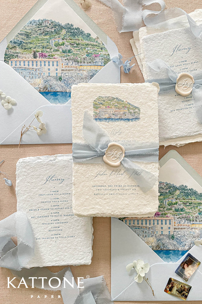 Luxury handmade paper wedding invitation suite featuring deckled edge cotton paper, custom watercolor Amalfi Coast venue artwork, dusty blue chiffon ribbon wraps, personalized wax seals, itinerary cards, and soft blue envelope liners.