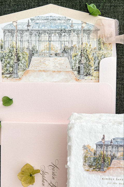 Detailed overhead photograph of a blush pink wedding envelope featuring a custom watercolor liner illustrating an elegant glass conservatory surrounded by soft greenery and landscaped gardens. The illustrated envelope is paired with coordinating blush pink stationery, personalized guest addressing, delicate fabric ribbon, and botanical accents on a dark green background.