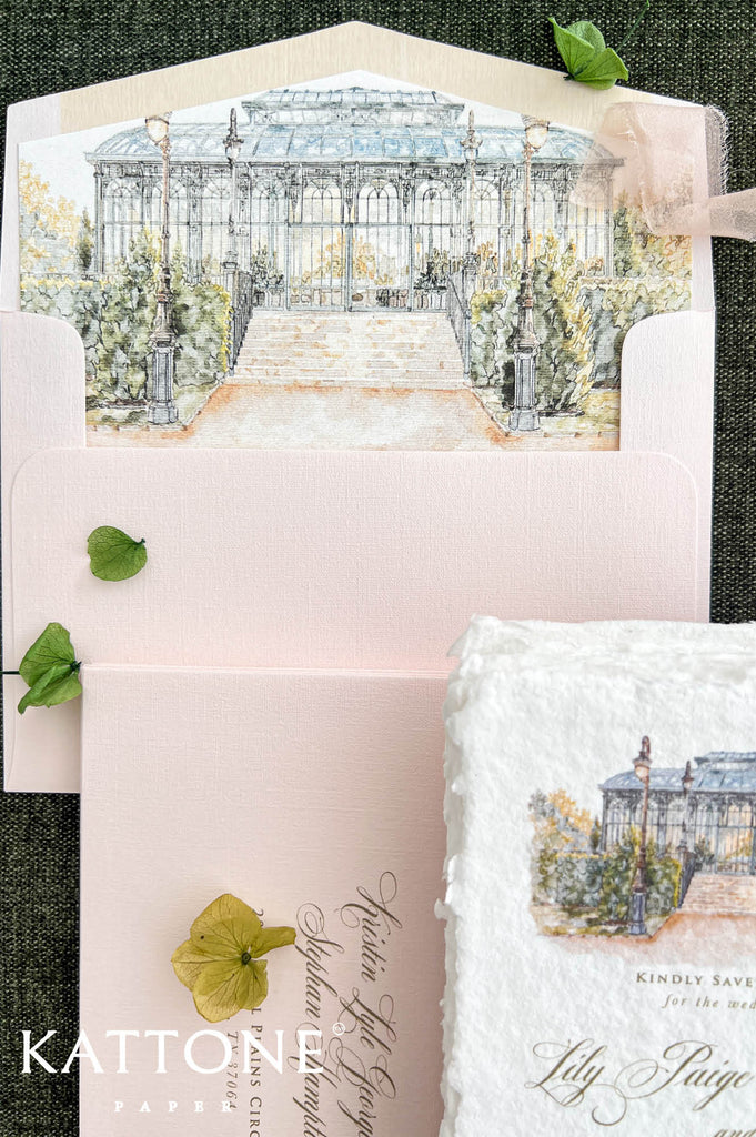 Detailed overhead photograph of a blush pink wedding envelope featuring a custom watercolor liner illustrating an elegant glass conservatory surrounded by soft greenery and landscaped gardens. The illustrated envelope is paired with coordinating blush pink stationery, personalized guest addressing, delicate fabric ribbon, and botanical accents on a dark green background.