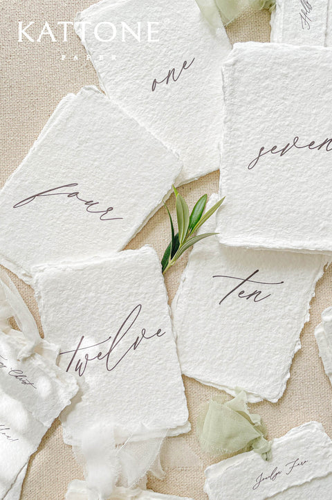 Handmade paper table numbers with handwritten script typography and deckled edges, fine art wedding decor.