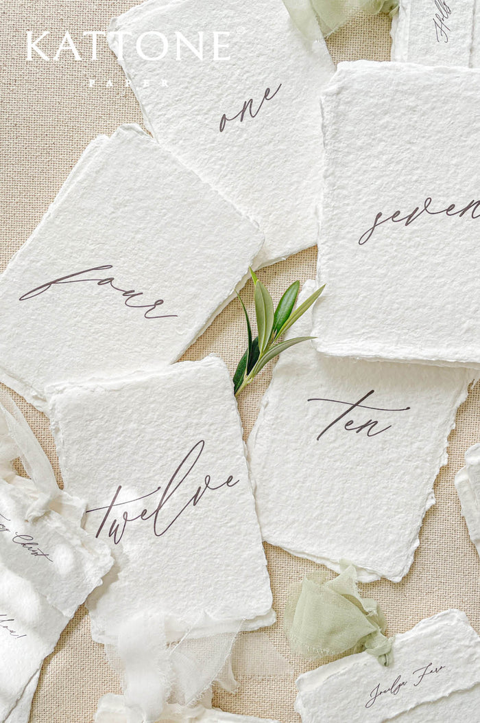 Handmade paper table numbers with handwritten script typography and deckled edges, fine art wedding decor.