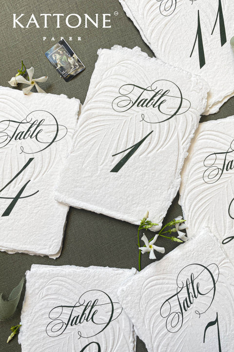Embossed handmade paper table numbers with deckle edges and elegant calligraphy, styled for a wedding reception.