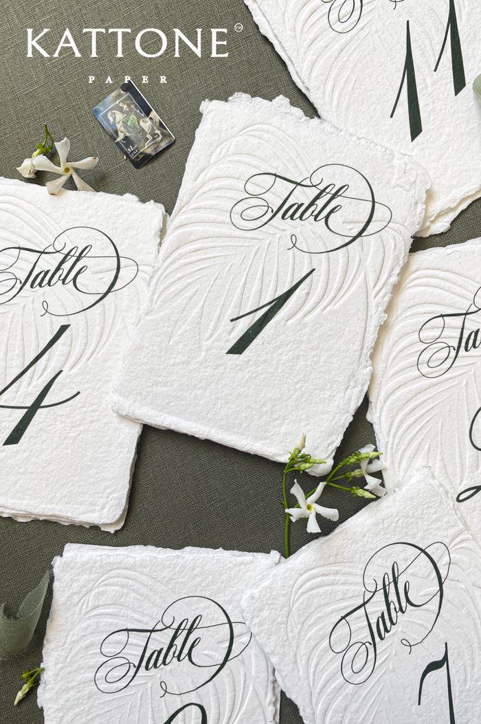 Embossed handmade paper table numbers with deckle edges and elegant calligraphy, styled for a wedding reception.