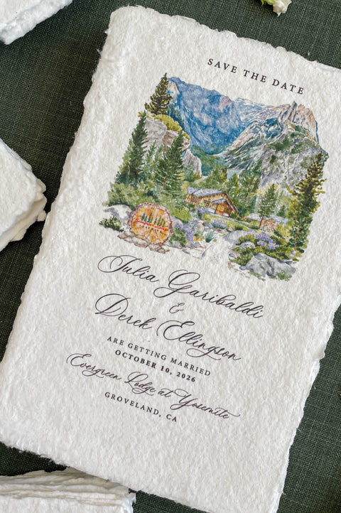 Close-up of handmade paper save the date card featuring a watercolor mountain wedding venue illustration.
