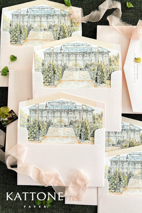Overhead flat lay of luxury blush pink wedding envelopes with custom watercolor liners depicting an elegant glass conservatory, grand stone staircase, vintage lampposts, and lush botanical greenery. Multiple open envelopes are arranged on a dark textured background alongside delicate blush silk ribbon, green leaves, and a coordinating envelope with personalized calligraphy address printing.