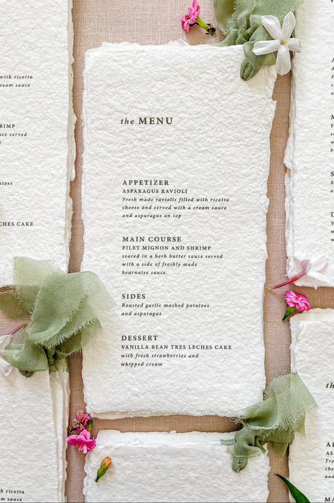 Handmade paper wedding menu with deckled edges, sage silk ribbon styling, minimalist typography, fine art reception stationery flat lay.