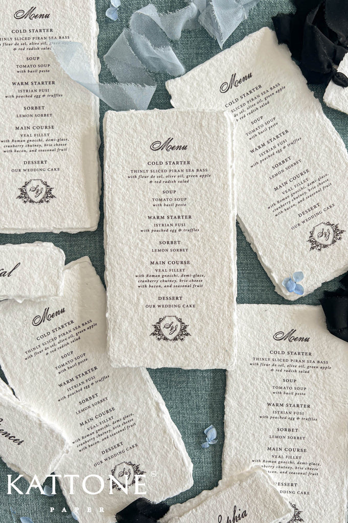 Overhead flat lay of luxury handmade paper wedding menu cards arranged on a muted blue linen background. Each ivory-white menu features naturally deckled edges, elegant black calligraphy, carefully centered dinner courses, and a personalized floral crest with the couple's initials. Soft dusty blue chiffon ribbon, black ribbon accents, and delicate blue flower petals complete the elegant wedding stationery arrangement.