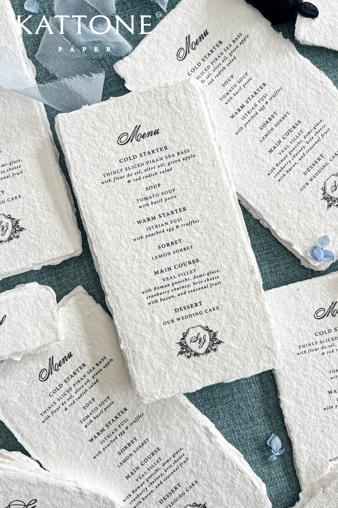 Angled overhead view of elegant ivory-white handmade paper wedding menus with naturally torn deckled edges, sophisticated black calligraphy, and a custom floral monogram crest. The central menu displays traditional serif typography with individual wedding dinner courses and an ornate botanical shield containing the couple's initials. Coordinating menus are styled with flowing dusty blue ribbon and delicate blue hydrangea petals on a textured blue background.